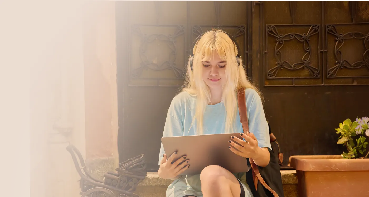Young woman with blonde hair wearing headphones and a light blue shirt using a tablet outdoors while sitting near a brown door and a potted plant.
