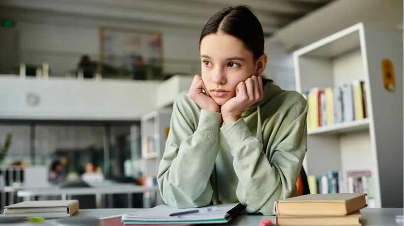 Teen girl in green hoodie sitting at a desk in a library, looking distracted with books and notebook in front of her.