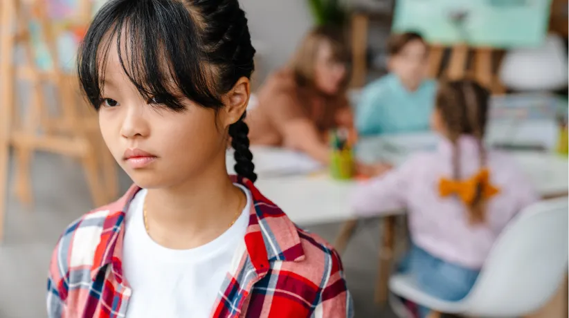 Young girl with braided hair and plaid shirt looking thoughtful, with other children studying in the background.