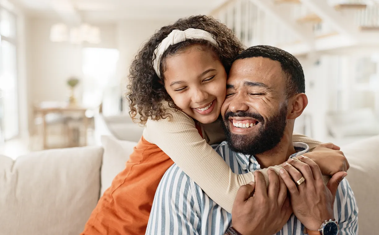 Young girl hugs a smiling man from behind on a beige couch in a bright living room.