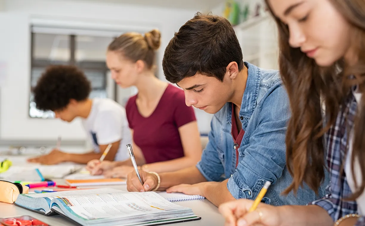 Four students sitting side by side at a desk, focused on writing in notebooks with an open textbook in front.