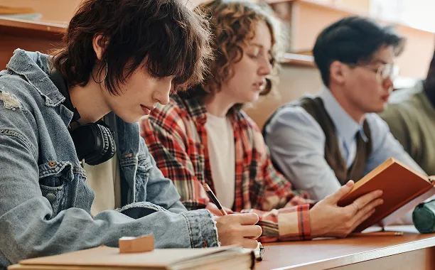 Three students sitting in a classroom, writing and reading attentively at their desks.