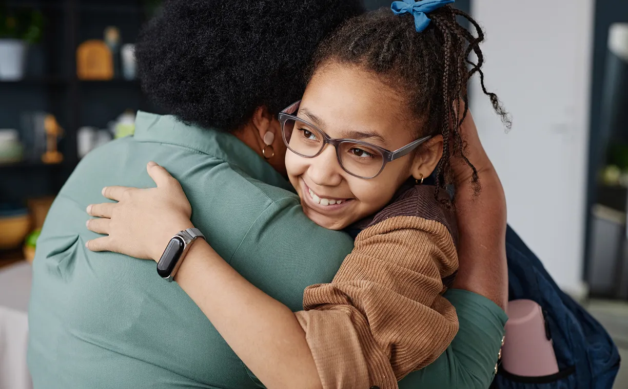 Smiling girl with glasses hugging an adult wearing a green shirt in a cozy indoor setting.