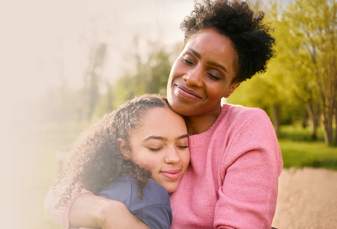 Woman in pink sweater hugging and smiling with a girl with curly hair outdoors in a park.
