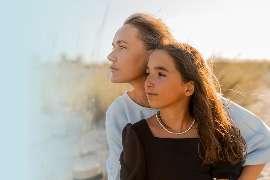 Curly woman in light blue top embracing her daughter in black dress with pearl necklace, both looking contemplatively into the distance at the beach.