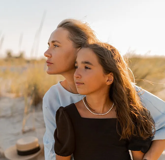 Curly woman in light blue top embracing her daughter in black dress with pearl necklace, both looking contemplatively into the distance at the beach.