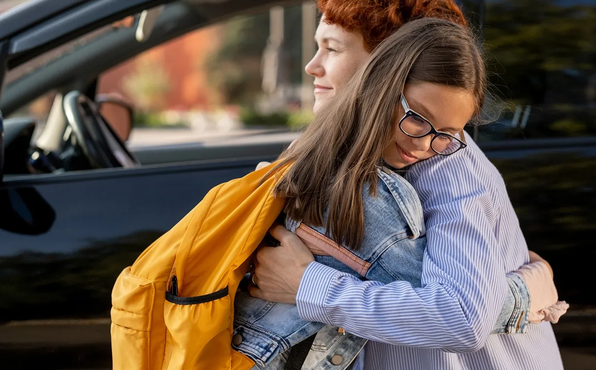 Smiling girl with glasses hugging an adult wearing a green shirt in a cozy indoor setting.