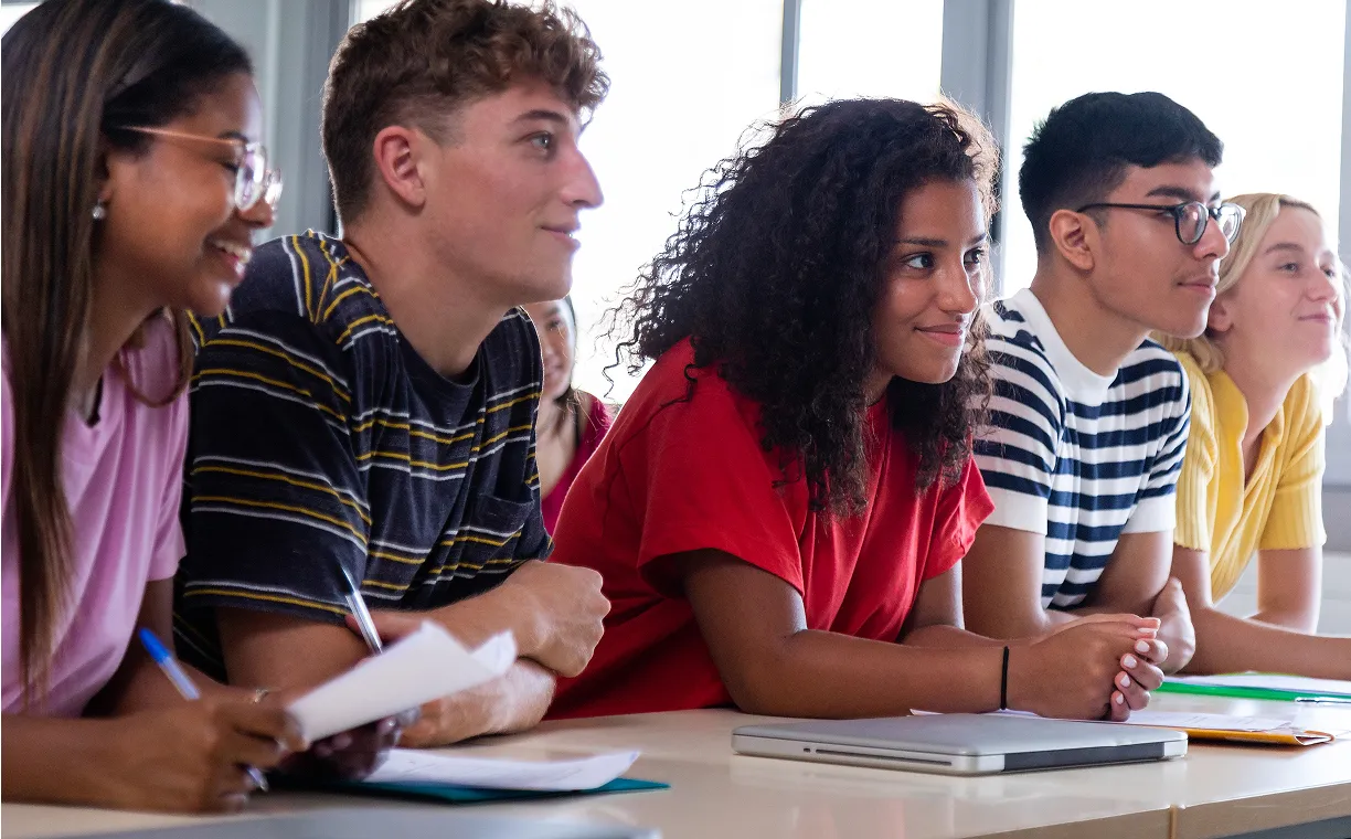 Three students sitting in a classroom, writing and reading attentively at their desks.