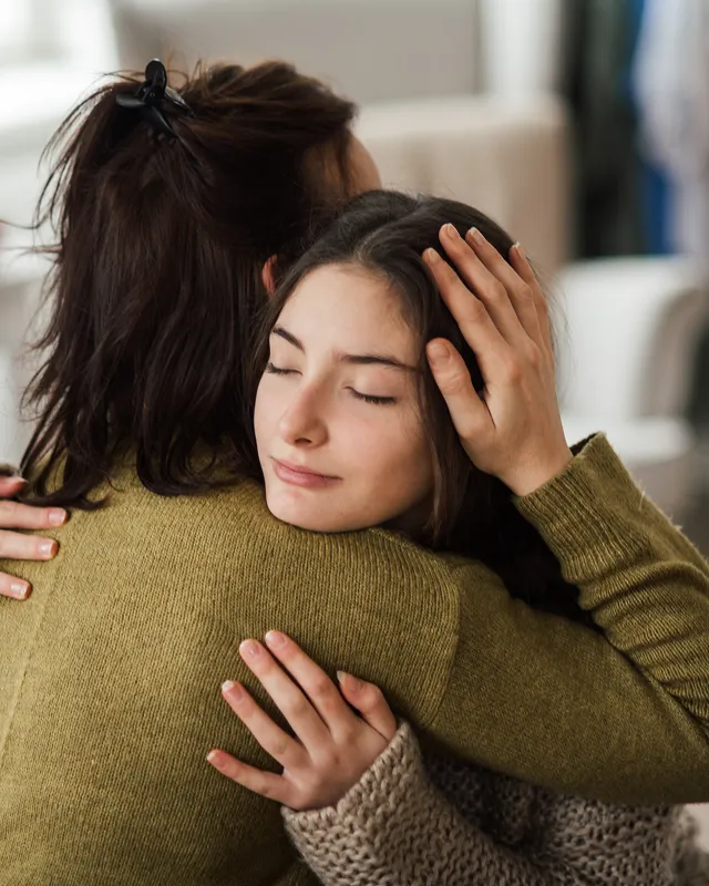 Two women hugging each other closely, one with eyes closed appearing comforted.