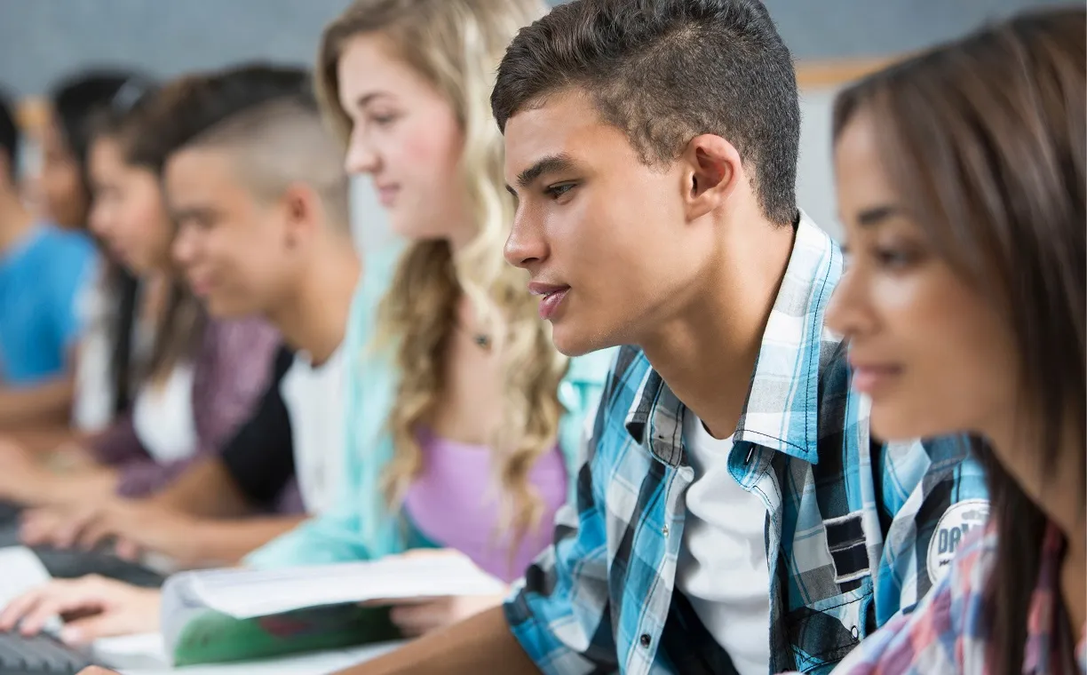 Three students sitting in a classroom, writing and reading attentively at their desks.