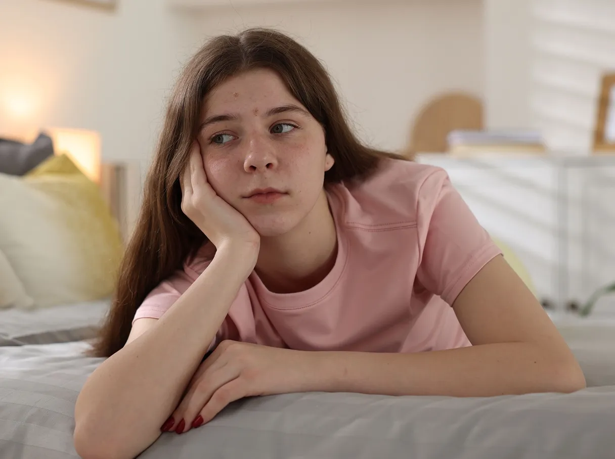Young person with short hair and glasses sitting cross-legged on a couch, looking thoughtfully downward.