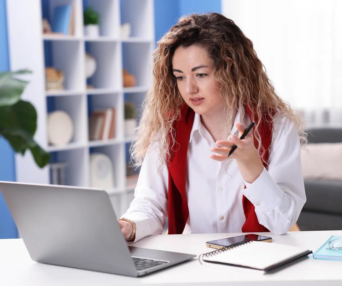 Woman with curly hair wearing a white shirt and red vest sitting at a desk, gesturing with a pen while looking at a laptop.