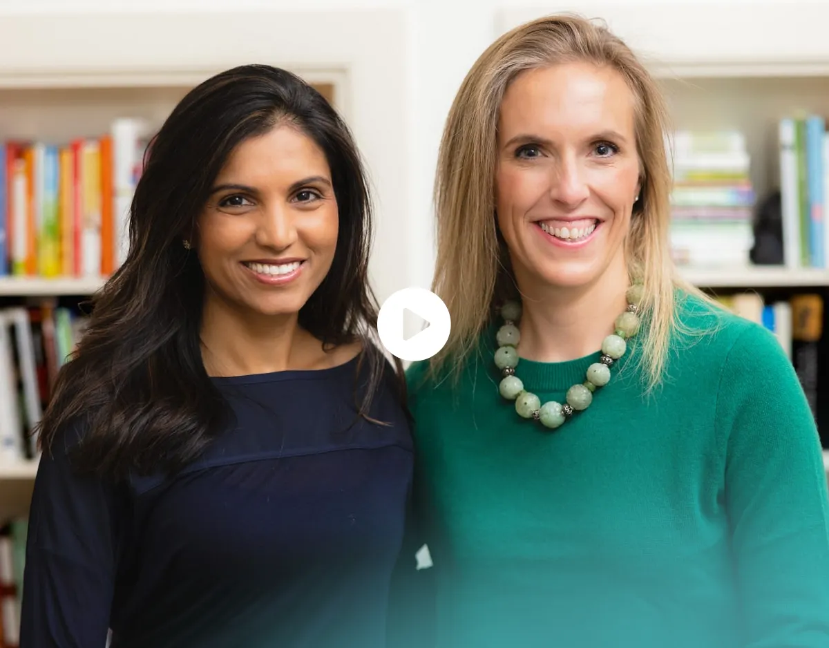 Two smiling women standing side by side in front of bookshelves, one with long dark hair wearing a navy top, the other with blonde hair wearing a green sweater and beaded necklace.
