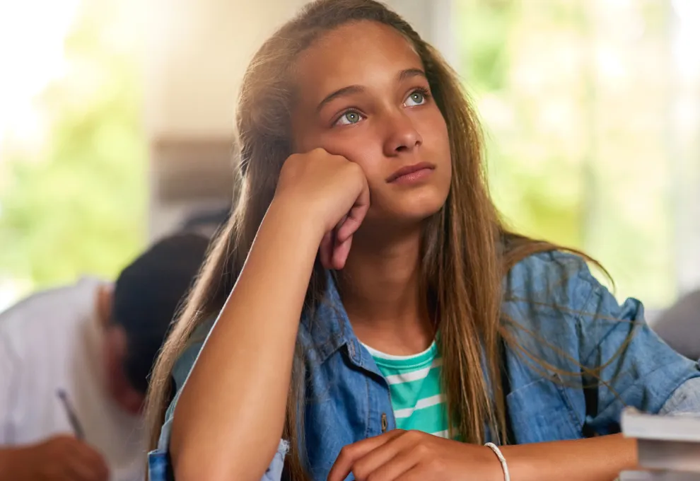 Thoughtful teenage girl with long hair resting her head on her hand in a classroom setting.