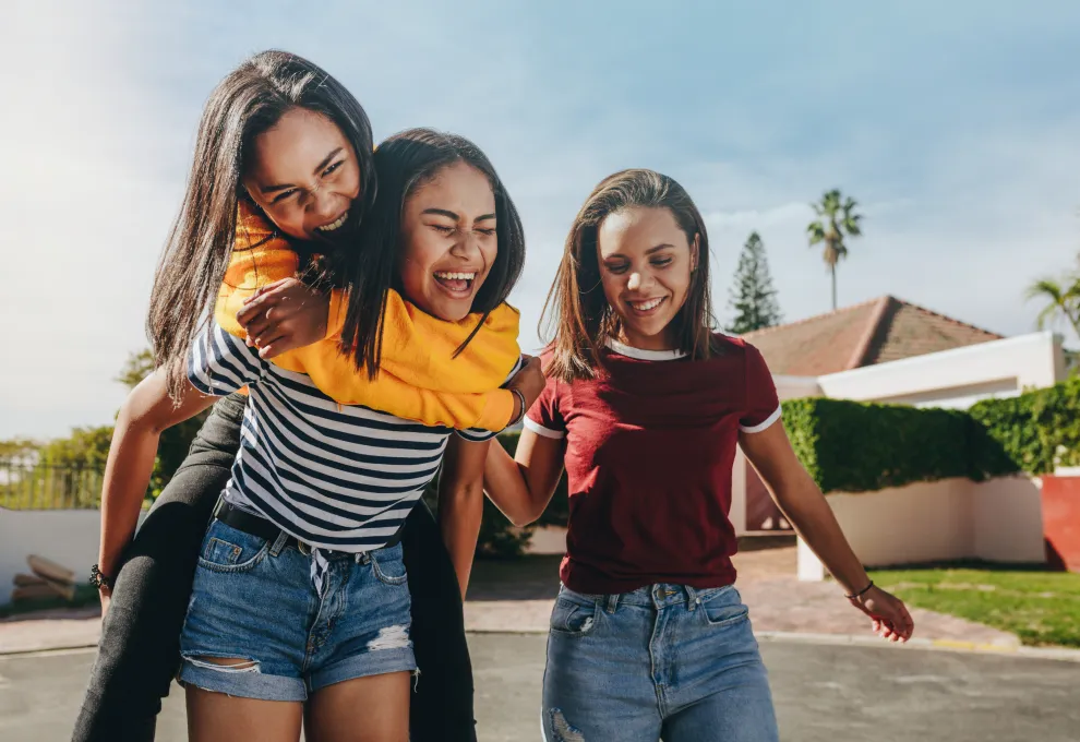 Three young women outdoors smiling and having fun, with one giving a piggyback ride.