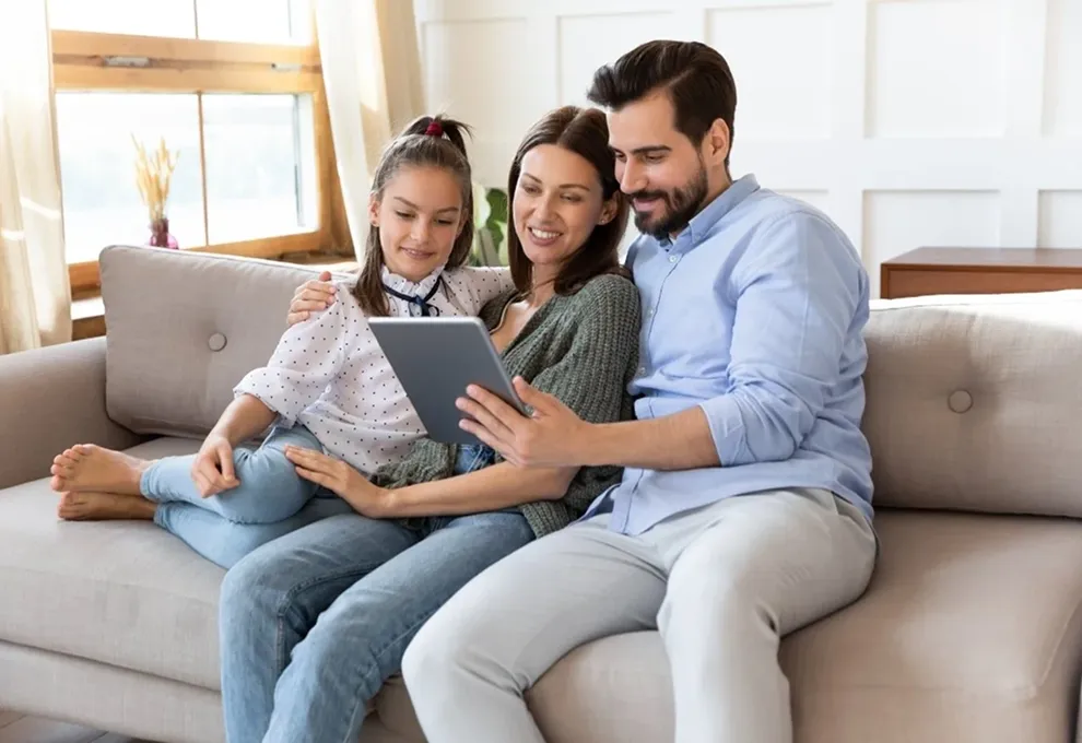Smiling family of three sitting on a couch looking at a tablet together in a bright living room.