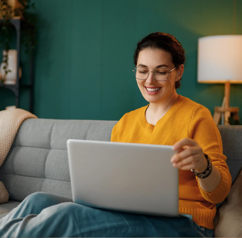 Smiling woman in glasses and orange sweater sitting on a gray couch using a laptop.
