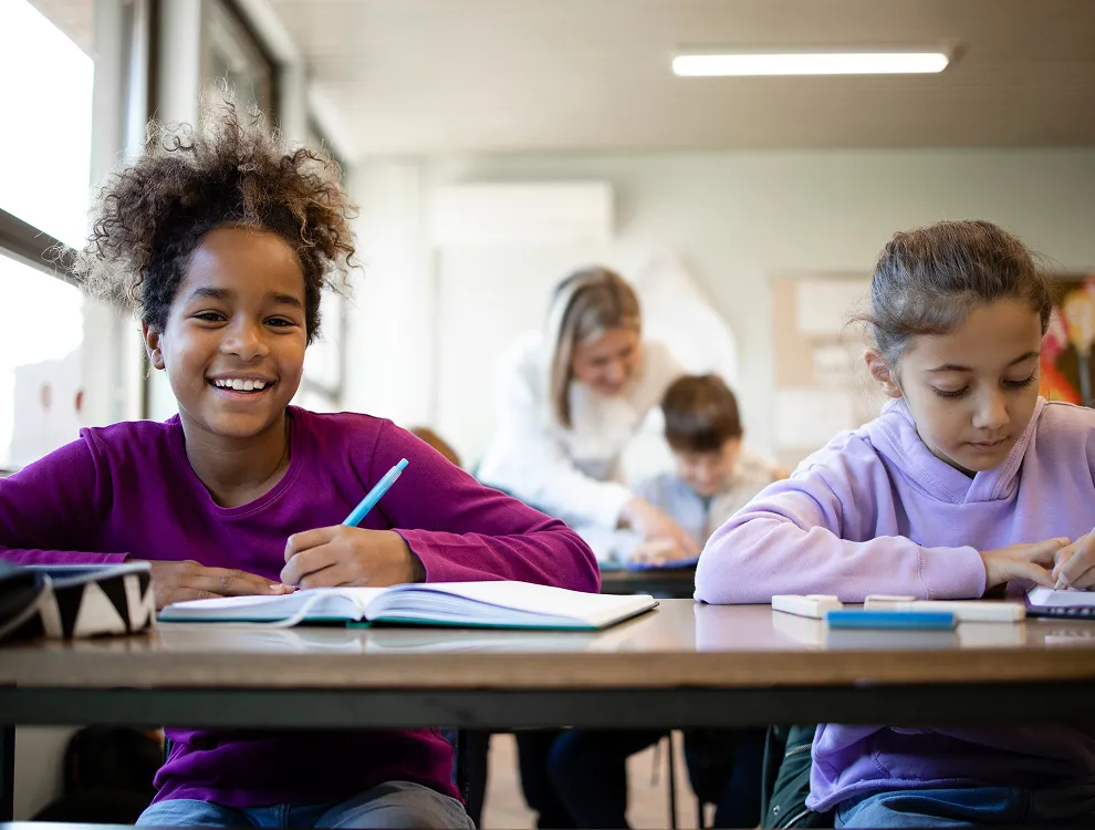 Two schoolchildren sitting at desks writing in notebooks with a teacher assisting students in the background.