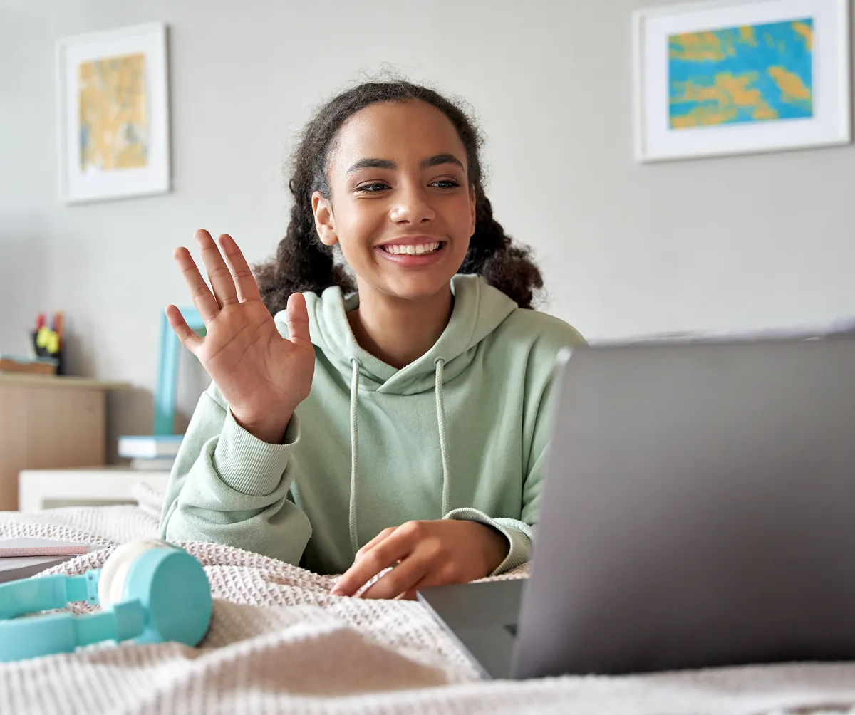 Smiling young woman in a light green hoodie waving during a video call on a laptop.