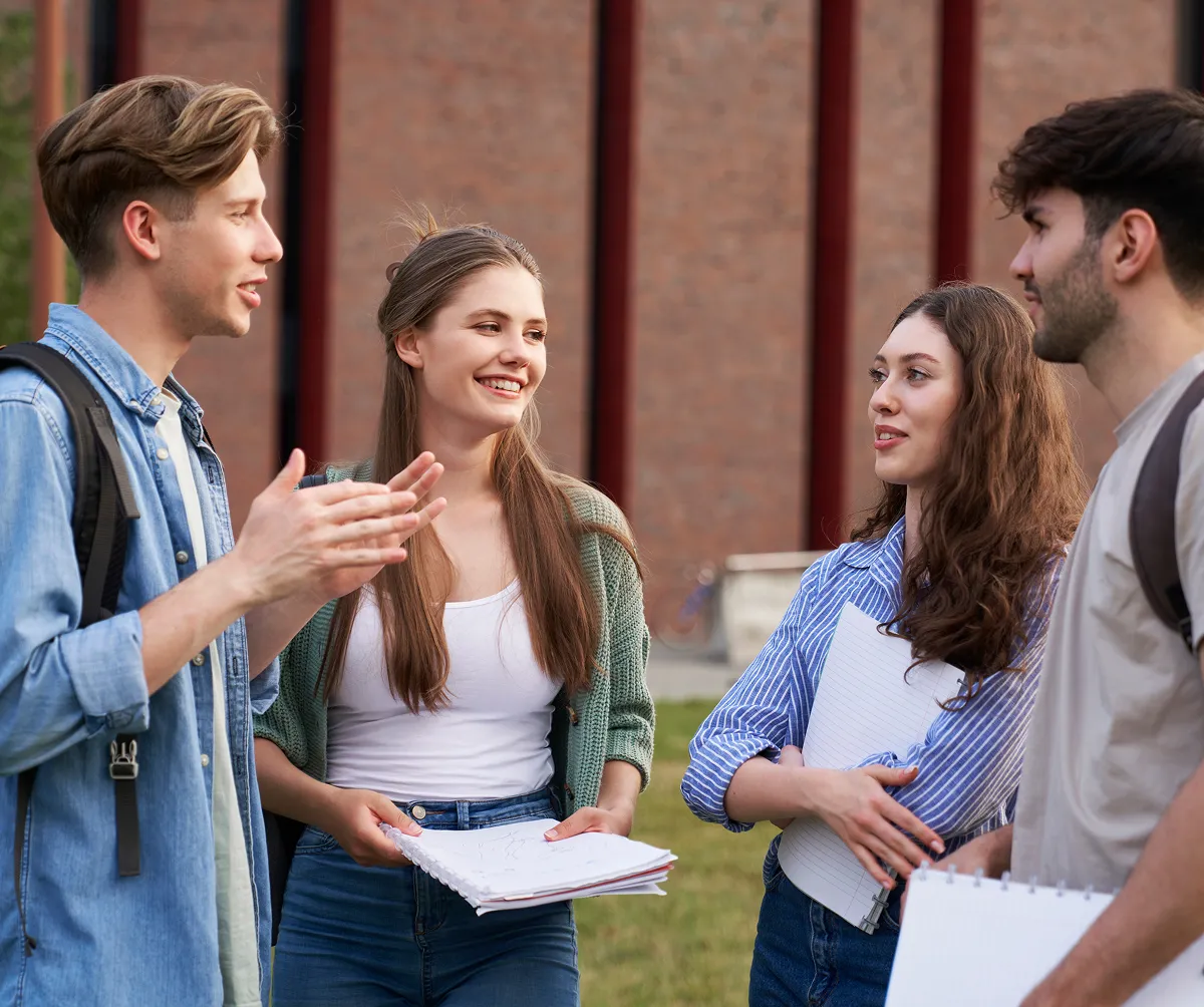 Four college students standing outside near a brick building, holding notebooks and engaging in conversation.