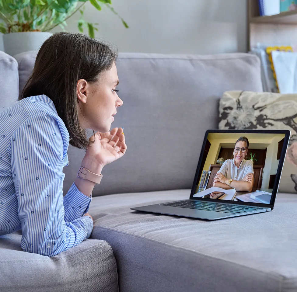 Woman lying on a couch engaging in a video call with a smiling professional on a laptop screen.