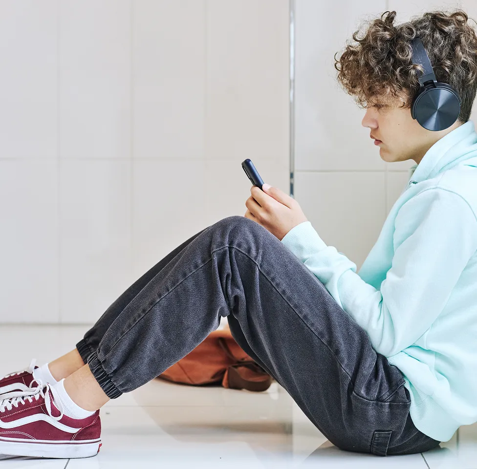Teenager with curly hair wearing black headphones, light blue hoodie, gray joggers, and red sneakers sitting on the floor while looking at a smartphone.