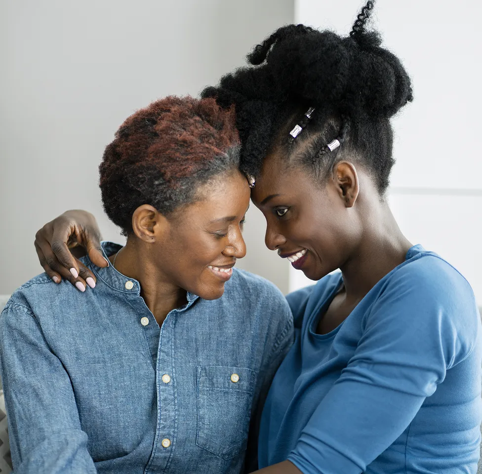 Two women leaning their foreheads together, smiling warmly, one with short curly hair and a denim shirt, and the other with styled natural hair and a blue top.