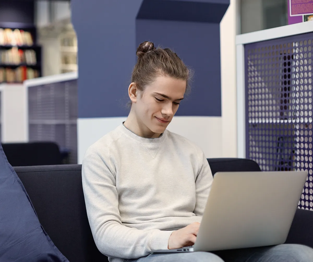 Young man with brown hair tied in a bun sitting on a couch, working on a laptop inside a modern library or study area.