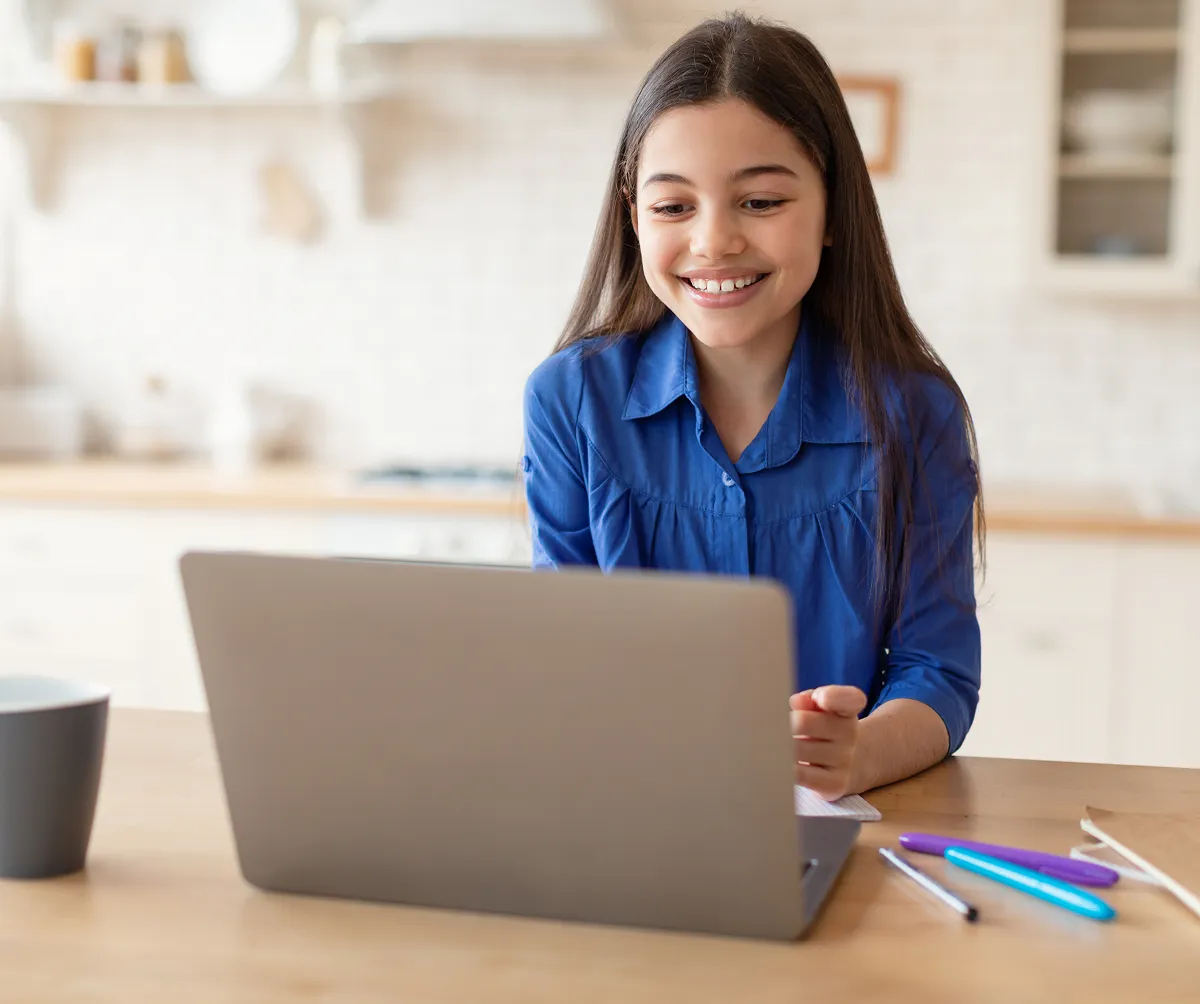 Smiling girl in a blue shirt using a laptop at a kitchen table with pens and a cup nearby.