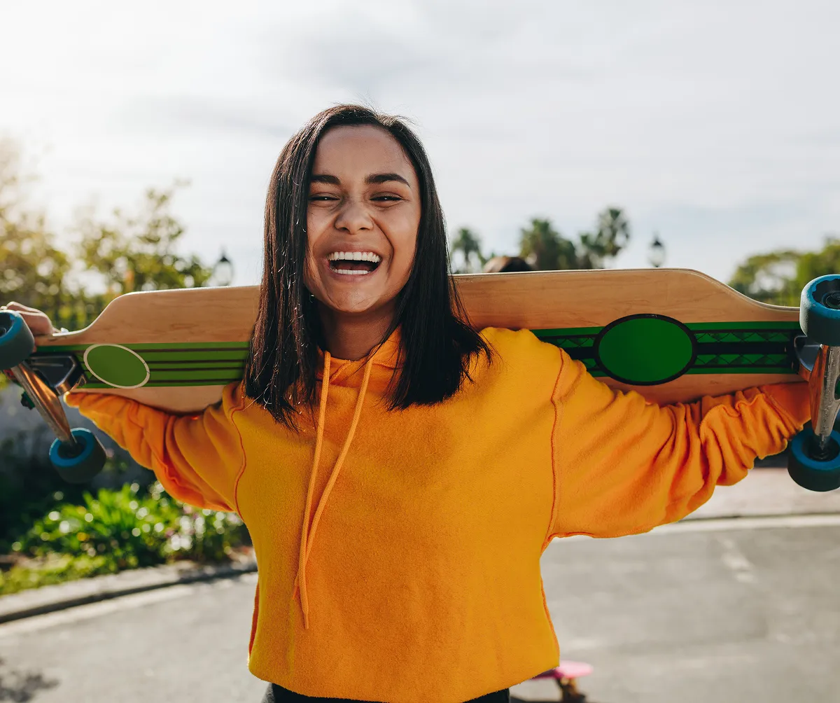 Smiling woman in an orange hoodie holding a longboard skateboard behind her shoulders outdoors.