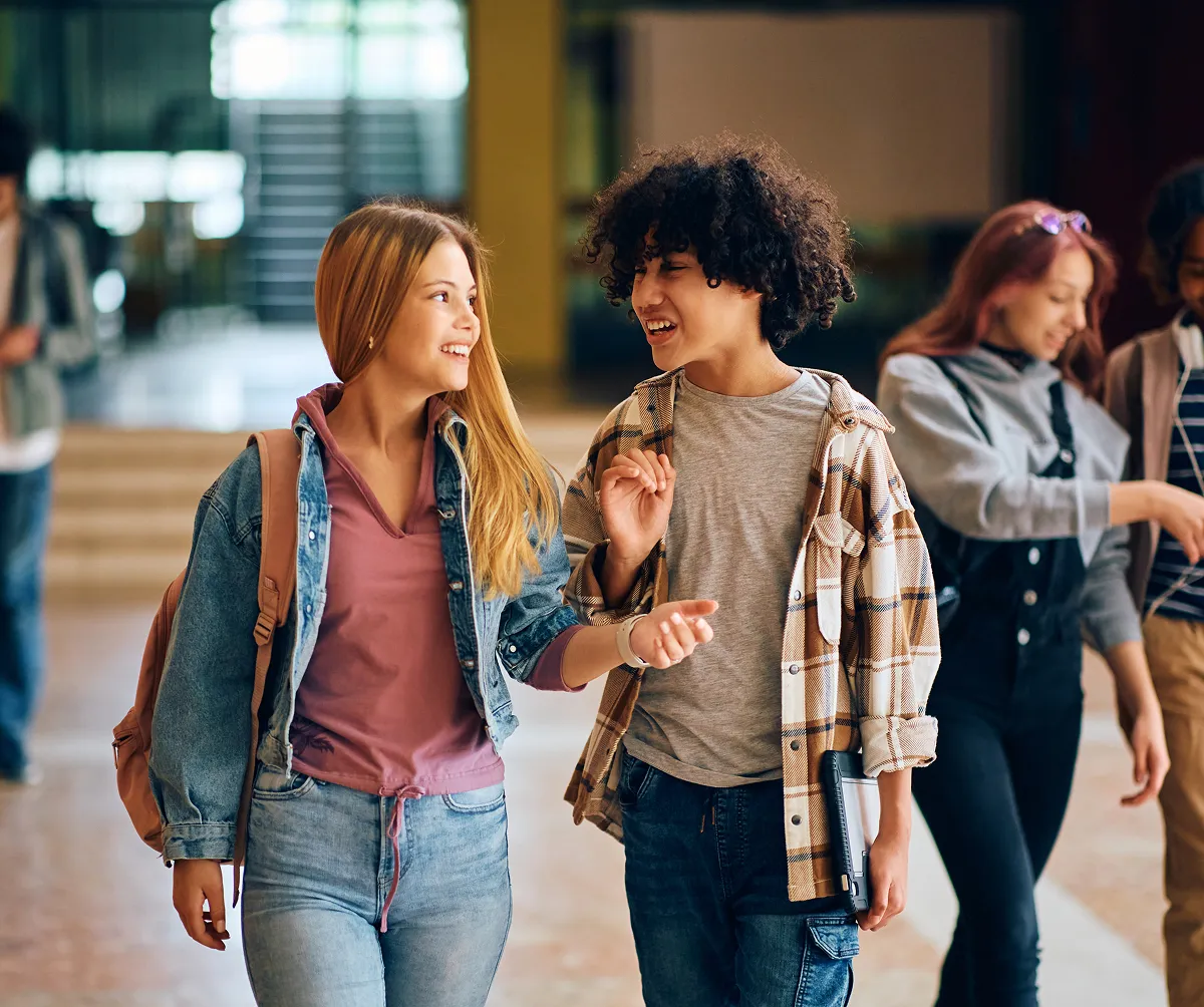 Two teenage students, a girl with long blonde hair and a boy with curly hair, smiling and talking while walking in a school hallway.