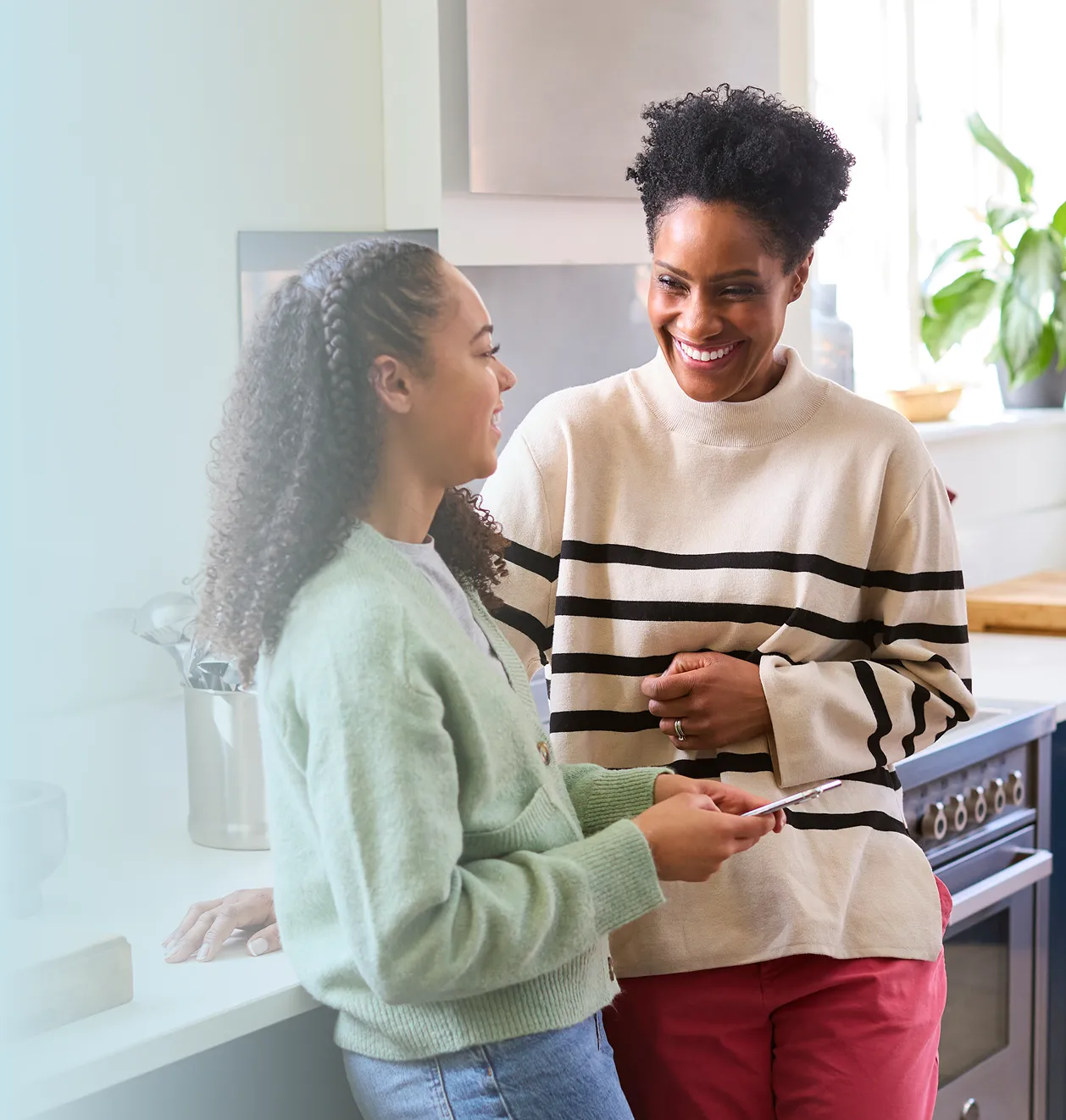 Two women smiling and talking in a kitchen, one wearing a striped sweater and the other holding a smartphone.