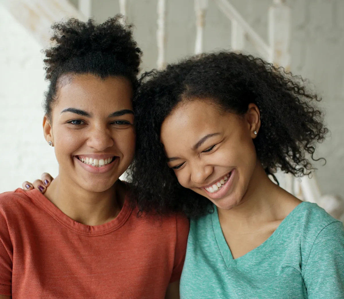 Two smiling young women with curly hair embracing, one wearing a red shirt and the other a green shirt.