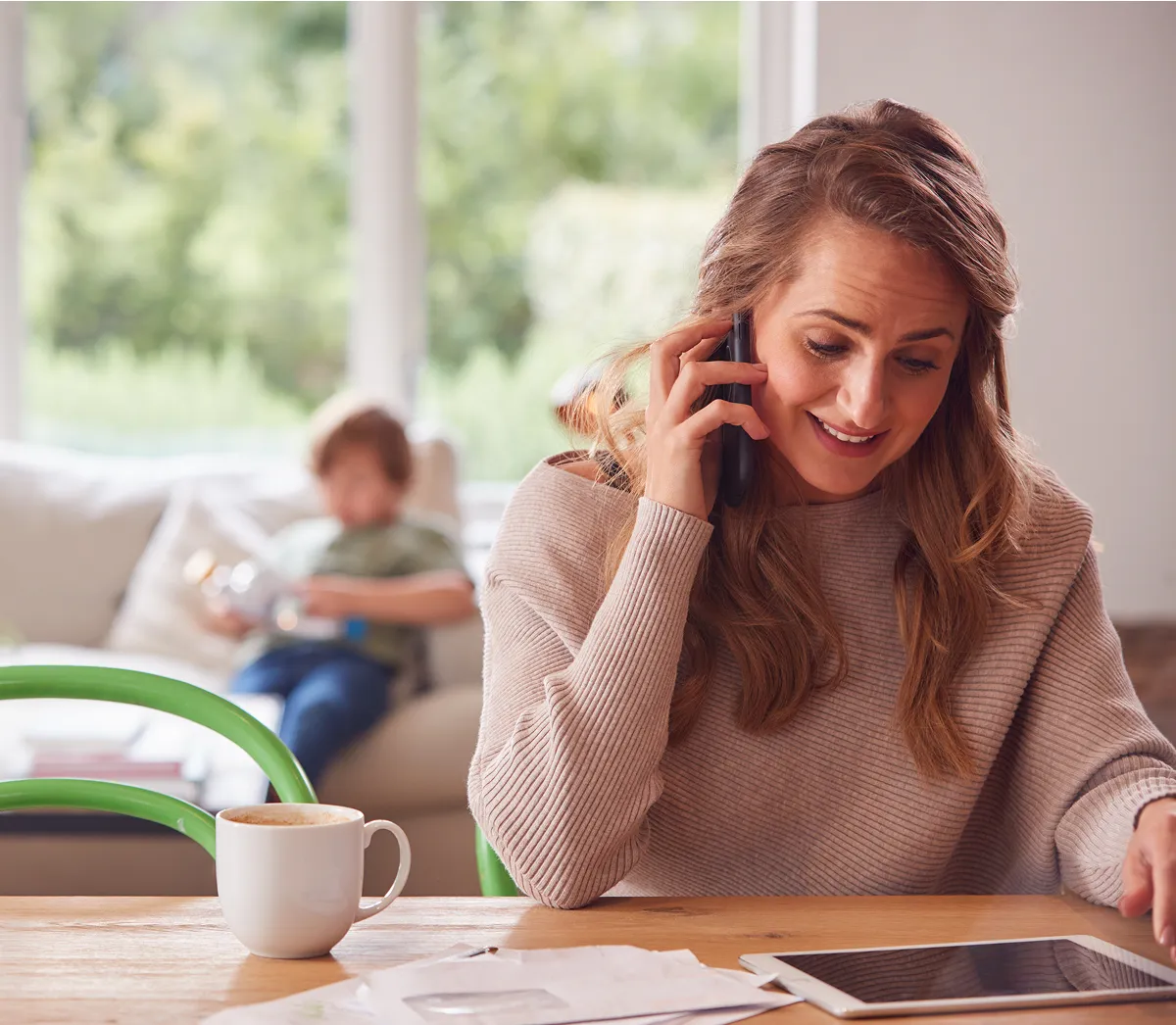 Smiling woman talking on the phone at a table with a cup of coffee, papers, and a tablet, with a child playing blurred in the background on a sofa.