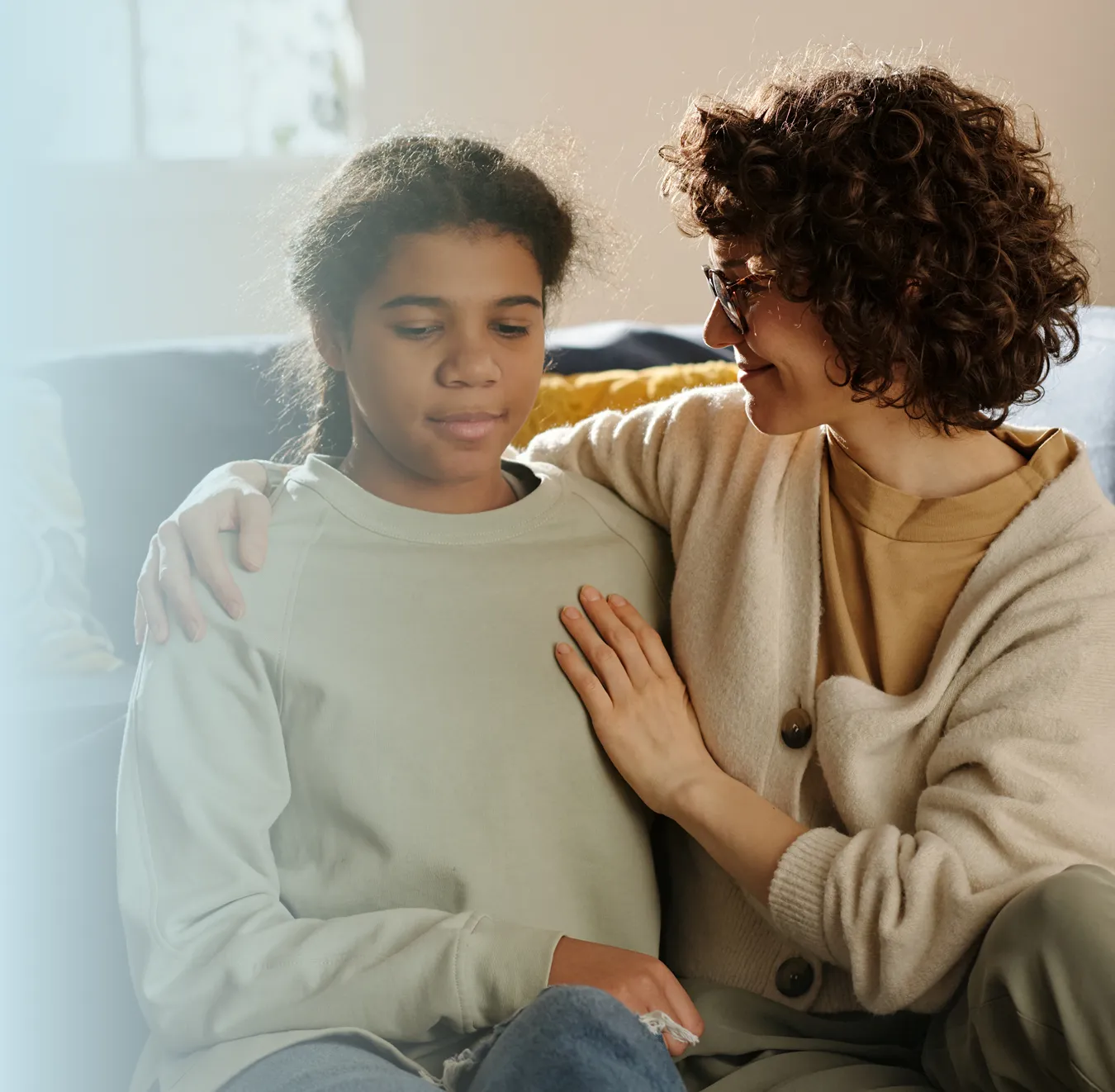 Woman with curly hair and glasses comforting a young girl on a couch by gently placing her hand on the girl's shoulder.