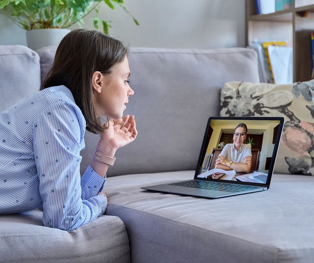 Woman lying on a couch having a video call with a smiling therapist on a laptop screen.