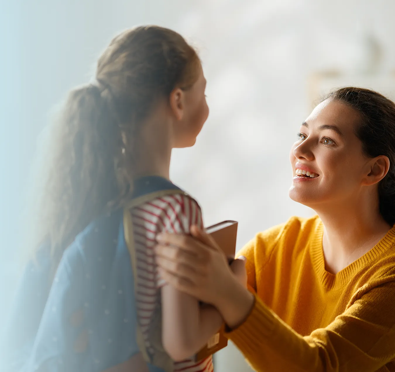 Smiling woman in yellow sweater holding the shoulders of a girl with a backpack and books, looking at each other.