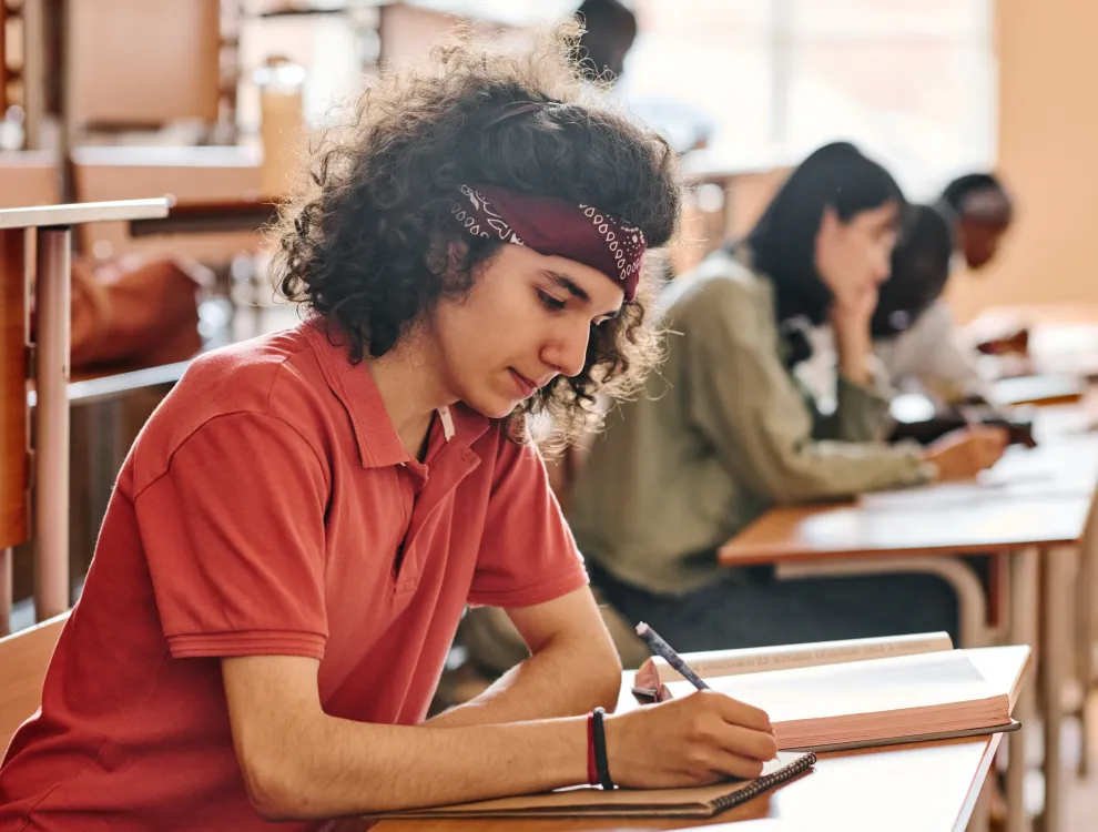 Student in red shirt and headband writing in notebook with open book on desk in classroom.