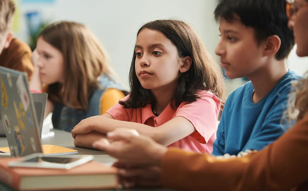 Three students sitting in a classroom, writing and reading attentively at their desks.