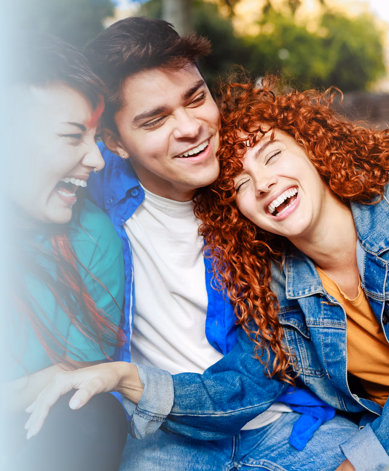 Three young friends laughing joyfully together outdoors, sitting close with arms around each other in warm sunlight.
