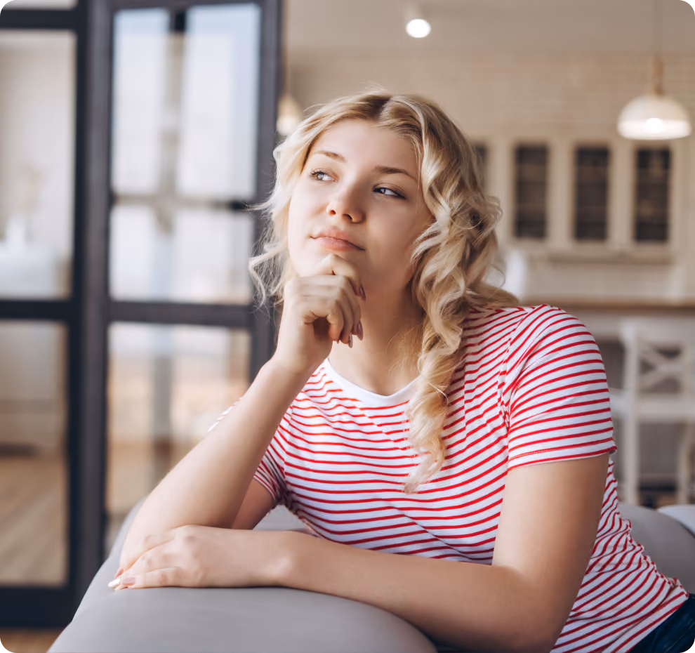 A young woman with curly blonde hair in a red-and-white striped shirt sits on a couch, resting her chin on her hand and gazing thoughtfully into the distance.
