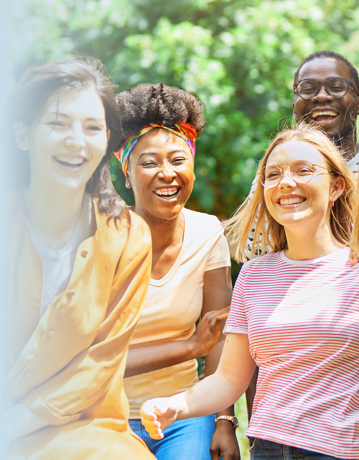 A group of four young adults laughing together outdoors in front of lush greenery on a sunny day, looking happy and relaxed.
