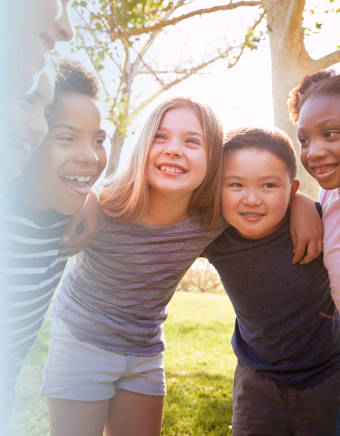 A group of smiling children stand close together with arms around each other outdoors in a sunny park, looking happy and full of energy.