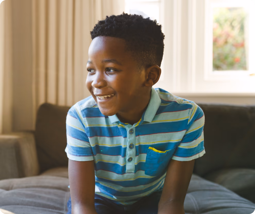 A young boy in a blue and green striped polo shirt sits on a couch, smiling and looking off to the side in a bright, cozy room.