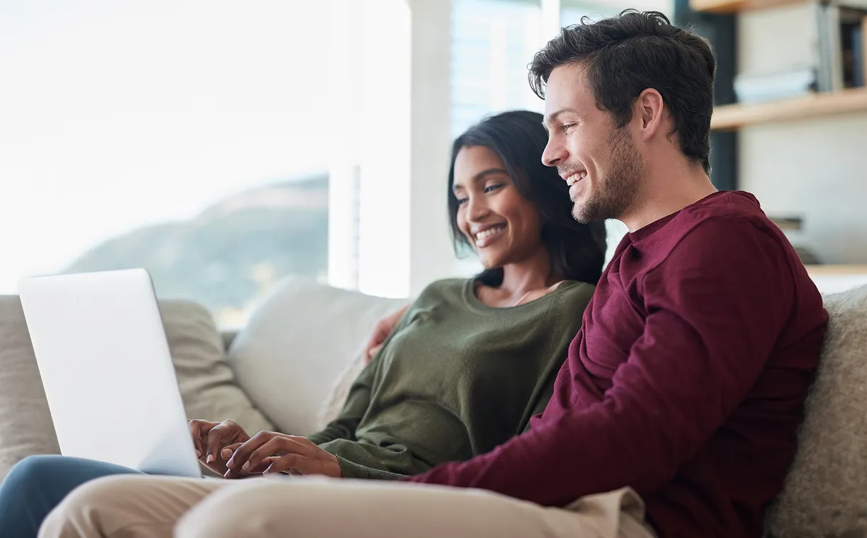 Smiling couple sitting closely on a sofa at home, sharing a laptop, creating a warm, relaxed, and connected atmosphere.