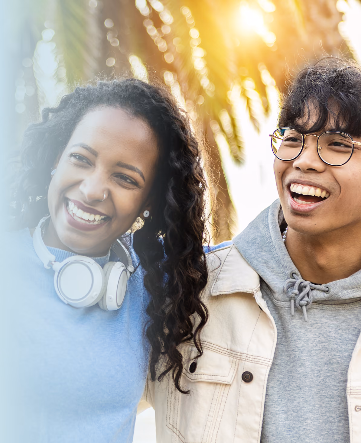 Two young people laughing together outdoors, one wearing white over-ear headphones around her neck and the other in glasses and a hooded jacket, with sunlit palm fronds behind them.