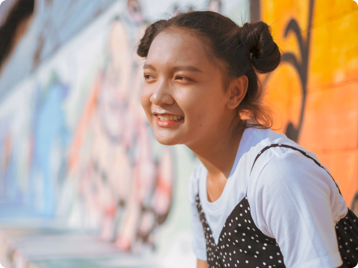 A smiling young woman with her hair in space buns, wearing a white t-shirt under a black polka-dot top, looking off to the side in front of a colorful mural.