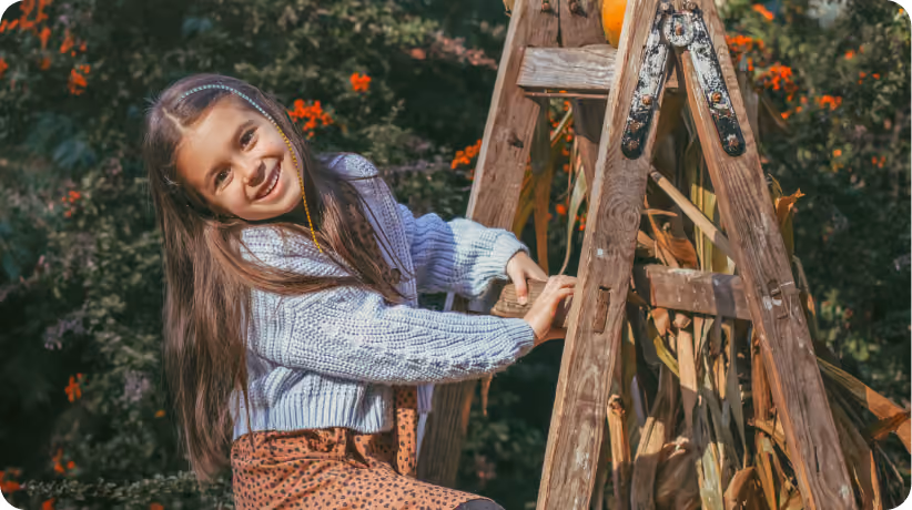 A smiling young girl in a light knit sweater and beaded hair strand climbs a wooden ladder outdoors, surrounded by autumn foliage and orange flowers.