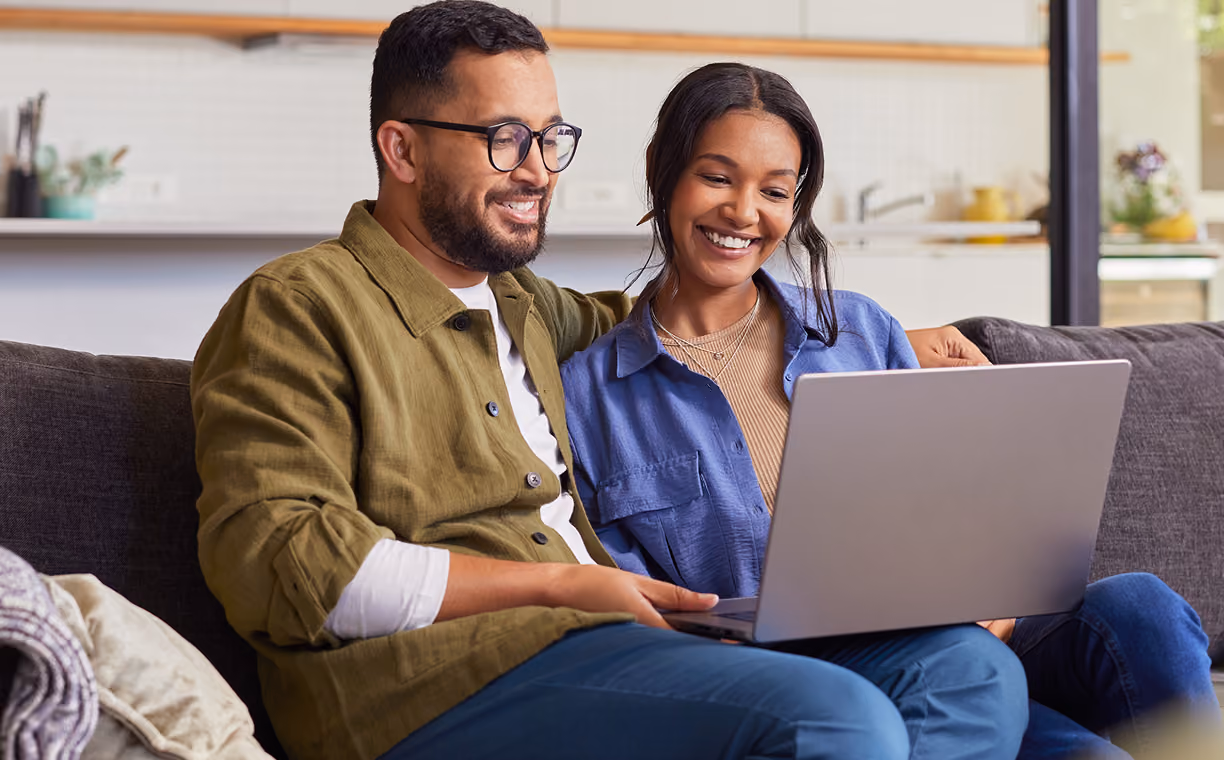 Smiling couple sitting closely on a sofa at home, sharing a laptop, creating a warm, relaxed, and connected atmosphere.