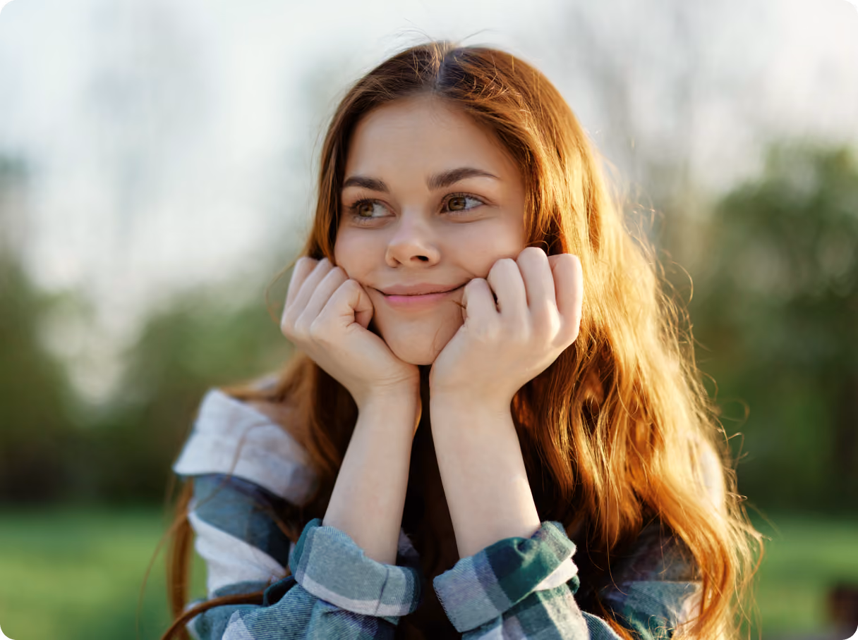 Young girl with long red hair resting her chin on her hands, smiling softly outdoors in a green natural setting.